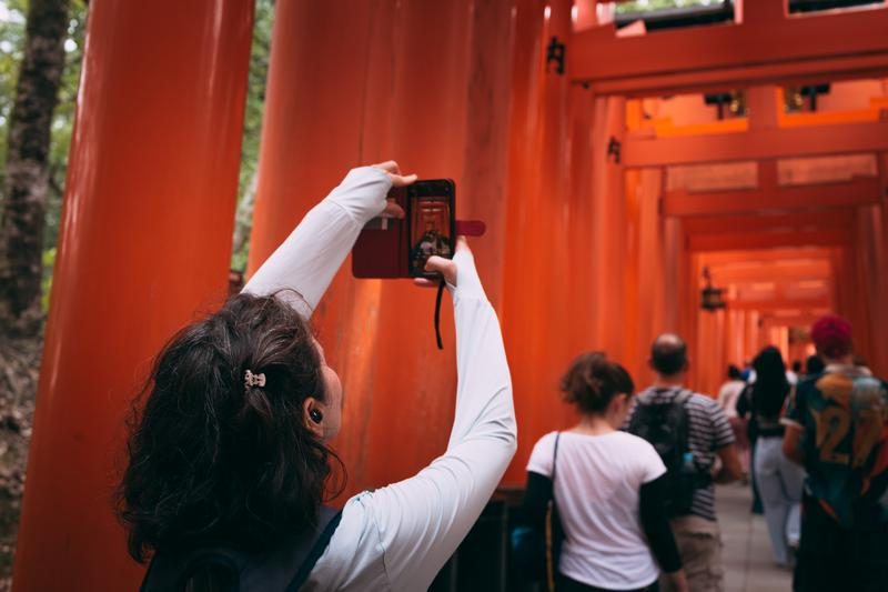 朱色の鳥居が並ぶ観光地をスマートフォンで撮影する人